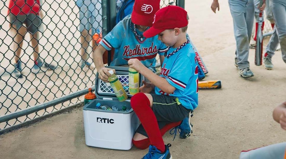 Two young Little League players take drinks from a white RTIC Road trip personal cooler filled with ice.