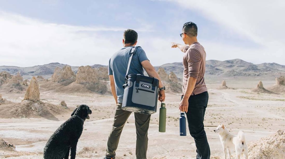 Two men and a dog hike desert mountains, one carrying a blue RTIC soft cooler over his shoulder with the strap.