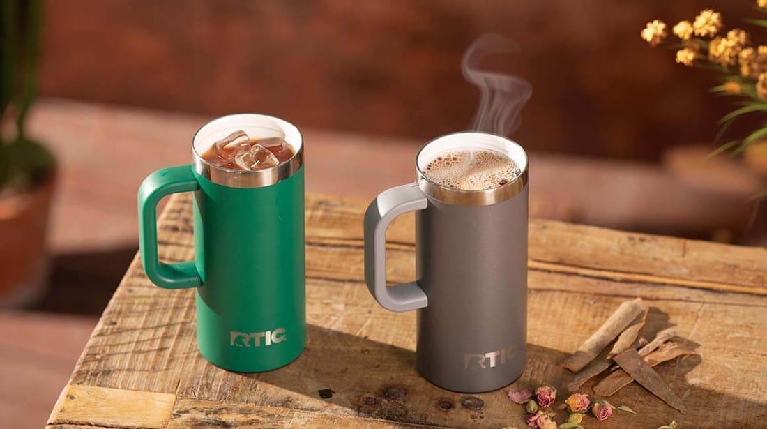 Green and gray RTIC Road Trip Travel Mugs displayed side by side on a wooden table.