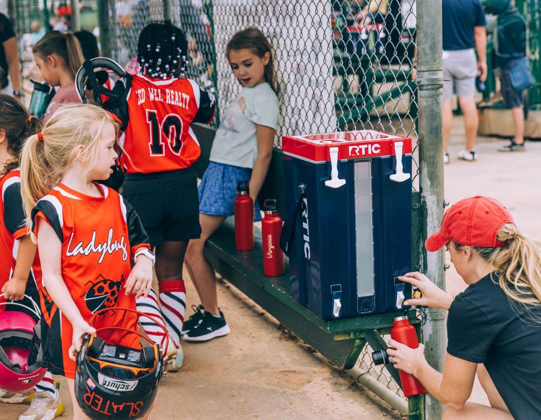 Mom fills her bottle from an RTIC Halftime Water Cooler beside girls in uniforms at a batting cage practice