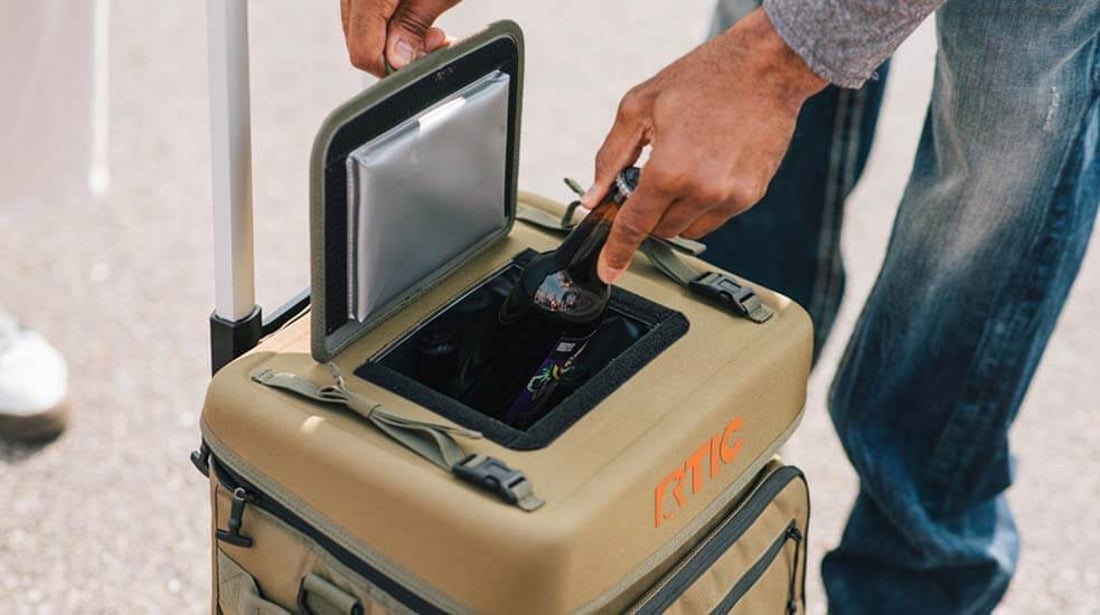 Man removes a drink from the top pocket of a green RTIC Everyday wheeled cooler.
