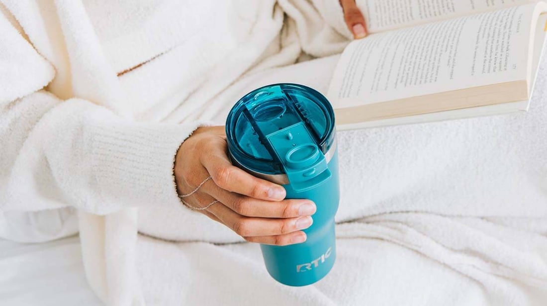 Woman reads a book while holding a blue RTIC Essential Tumbler.