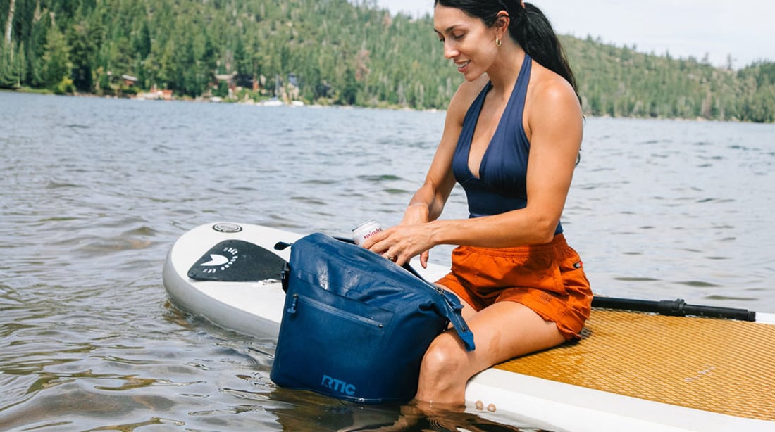 A woman sitting on a paddleboard with the RTIC Marina Waterproof Tote Cooler.