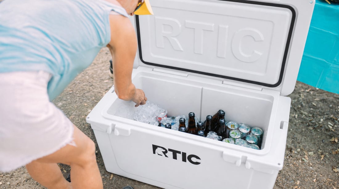 Person adding ice to an open RTIC cooler filled with bottled drinks and canned beverages.