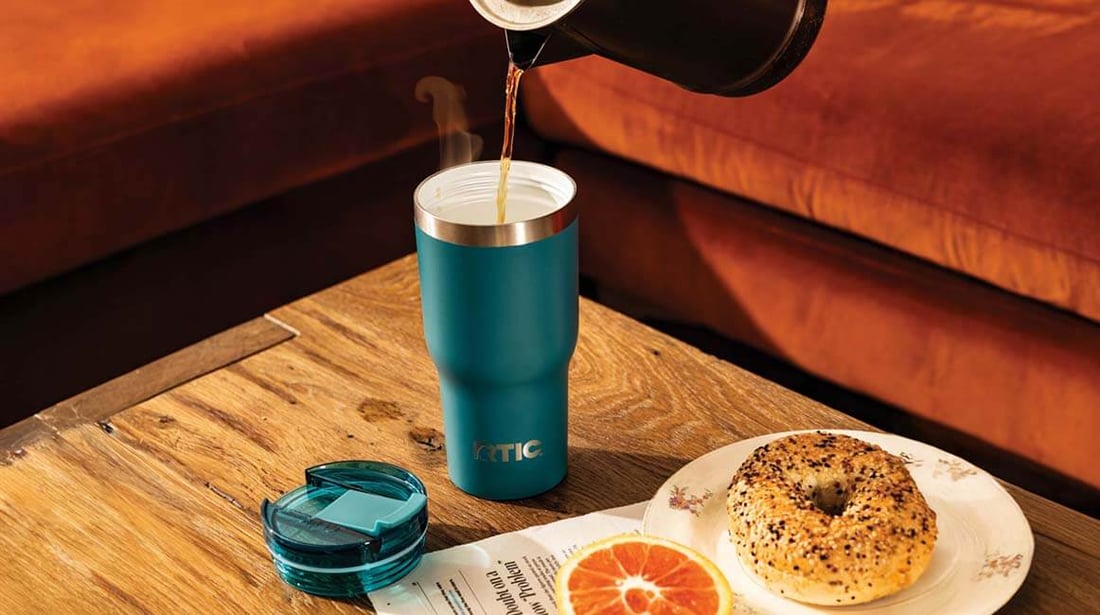Coffee poured into a blue RTIC Essential Tumbler on a table beside a bagel, with a couch in the background.