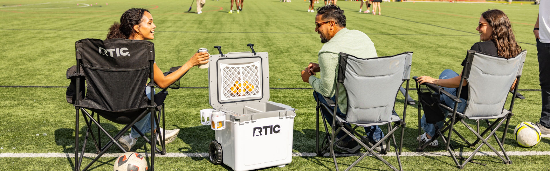 Friends on the sideline around a white RTIC wheeled cooler.