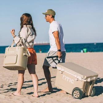 Two people walk on a sandy beach carrying a cooler bag and a wheeled cooler, with the ocean visible in the background.