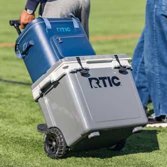Person pulling a grey RTIC wheeled cooler with a navy blue RTIC soft cooler stacked on it.
