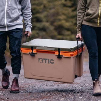 Two people carrying a tan RTIC hard cooler by the rope handles.