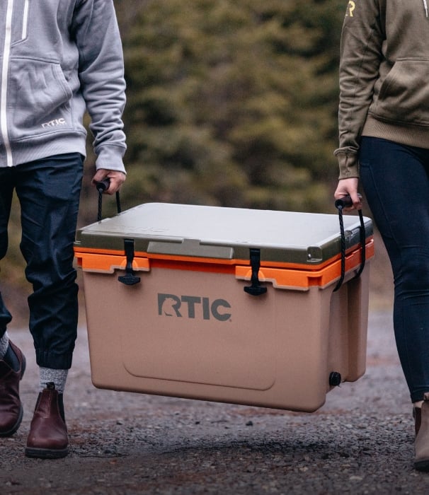 Two people carrying a tan RTIC hard cooler by the rope handles.