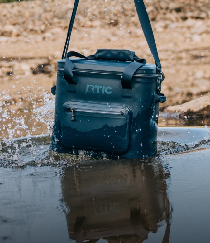 Black RTIC soft cooler sitting in shallow water with splashing water around it.