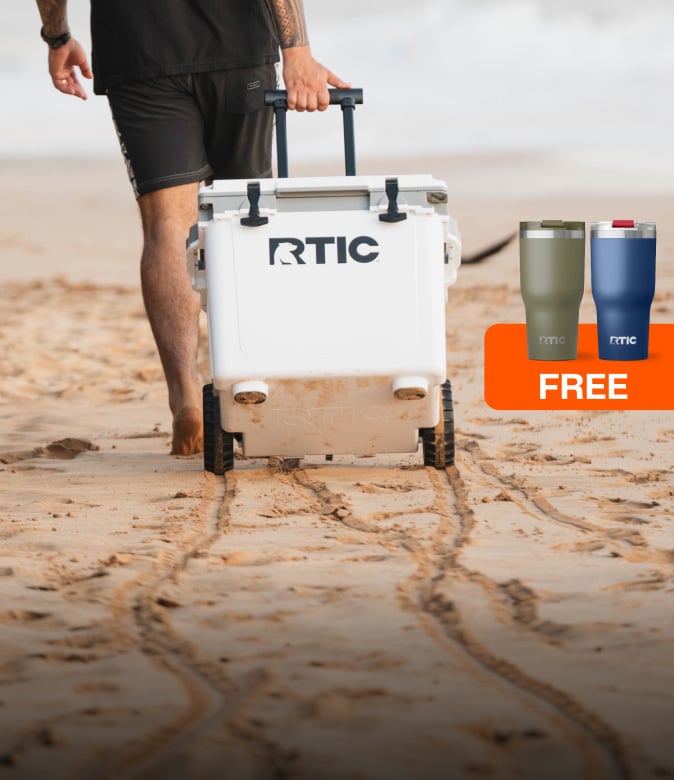 A person pulls a white RTIC wheeled cooler across a sandy beach, with two tumblers and a FREE promotional graphic.