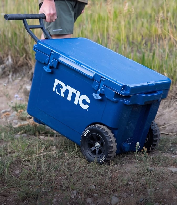 Person pulling a large blue RTIC cooler with wheels through a grassy outdoor area.