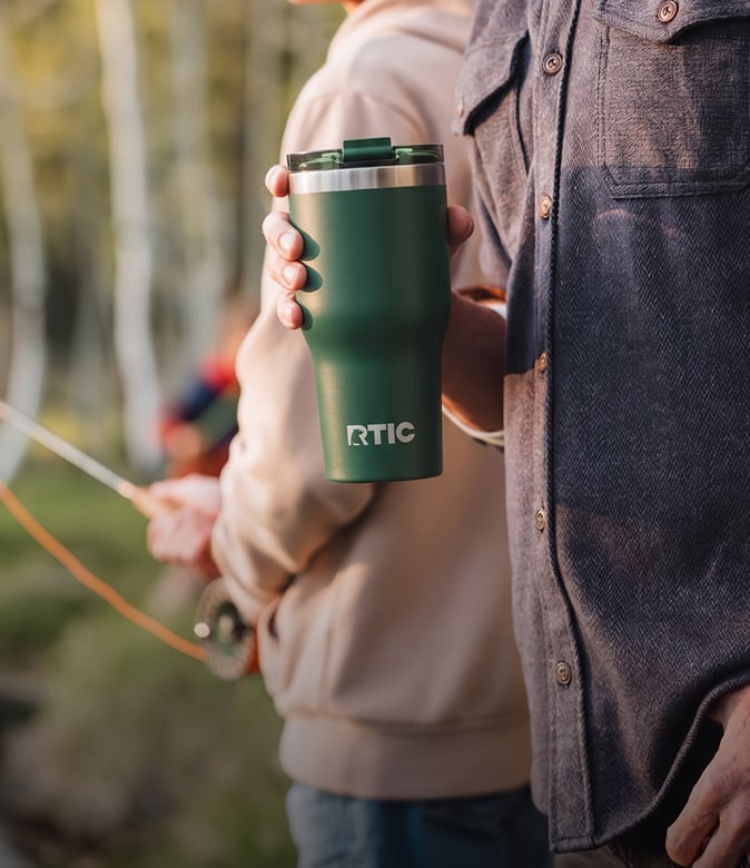 Man holding a green RTIC tumbler outdoors while another person fishes in the background.