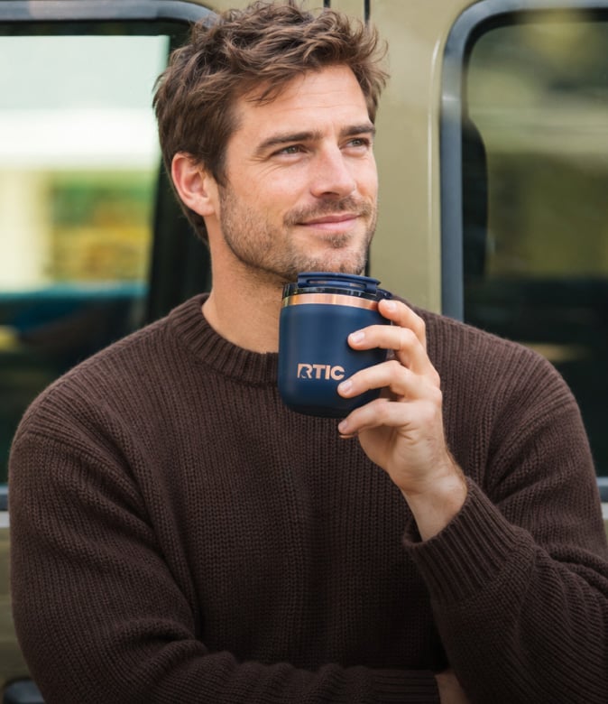 A man in a brown sweater standing outdoors and drinking from a dark blue RTIC insulated coffee cup.