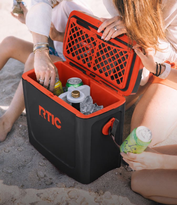 Woman opening a light black and orange RTIC cooler filled with ice and canned drinks on a sunny beach.