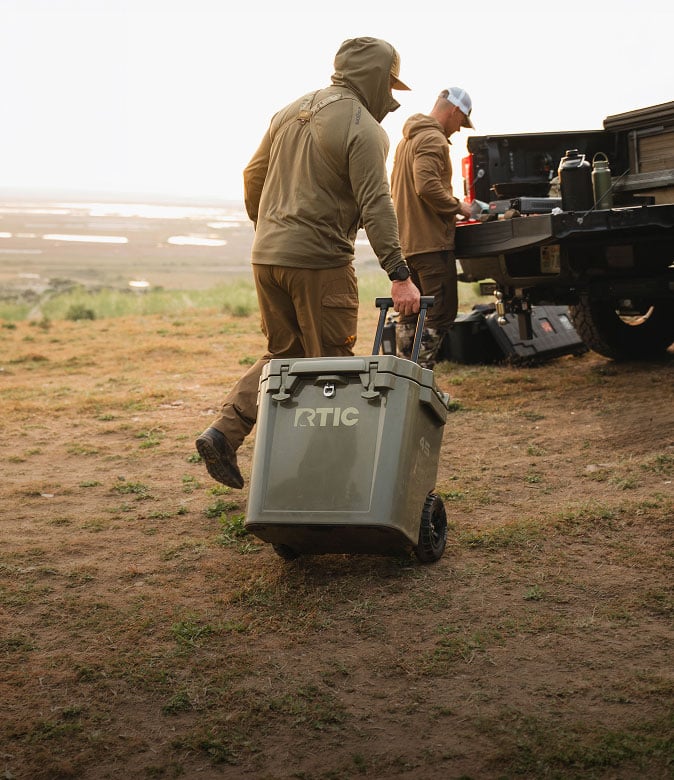 Two people outdoors near a pickup truck, with one pulling a green wheeled RTIC cooler across a grassy field.