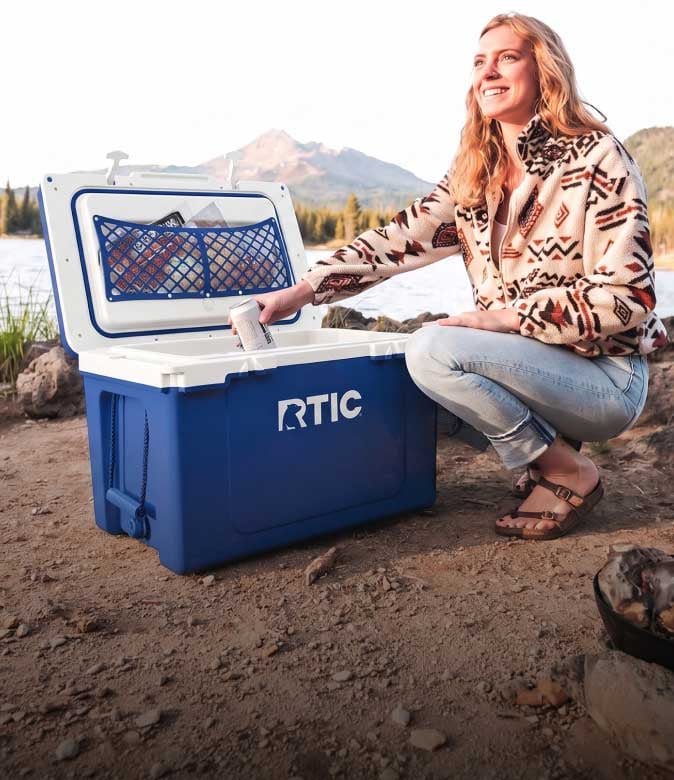 • A smiling woman kneels on the ground next to an open, blue RTIC hard-sided cooler and reaches for a drink inside.