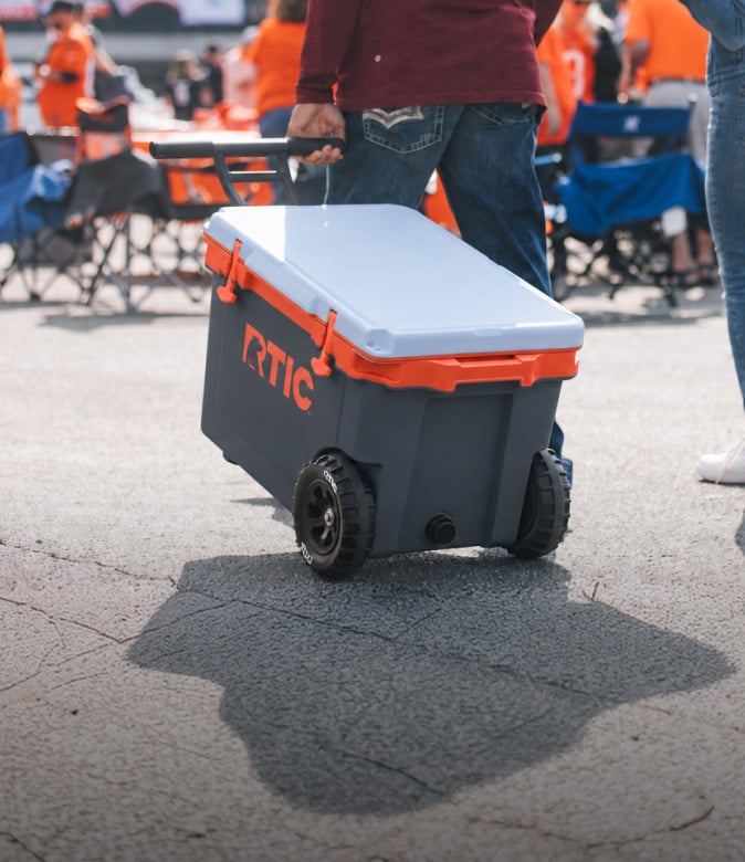 Person pulling a large light black wheeled RTIC cooler with a grey blue lid and orange trim.