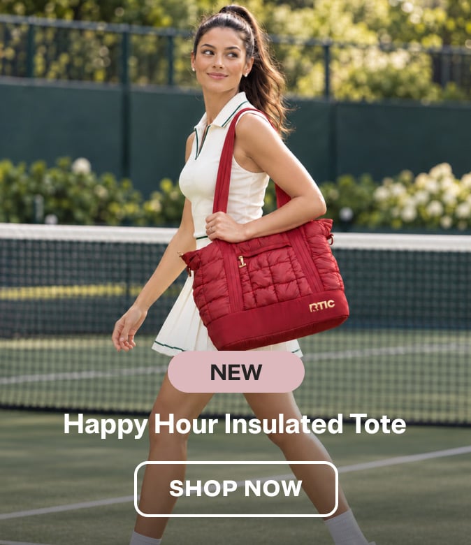 Woman in a white tennis dress carrying a black cherry RTIC quilted tote bag while walking on an outdoor tennis court.