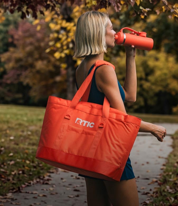 Woman drinking from an orange RTIC bottle while carrying an orange tote bag on a leafy autumn path.