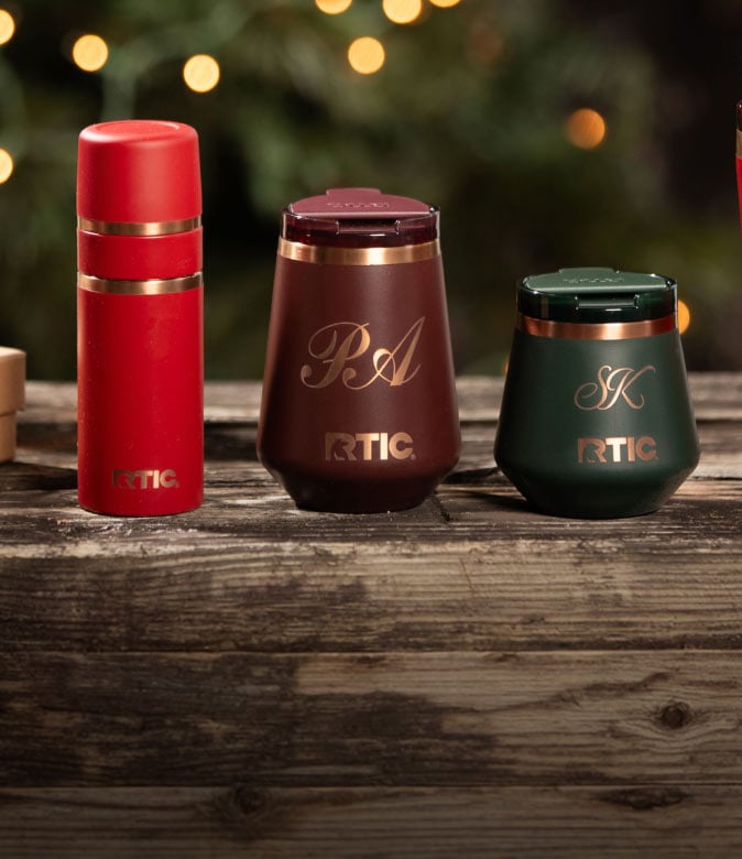 Red, brown, and green RTIC drinkware with gold accents and personalized monograms displayed on a rustic wooden table.
