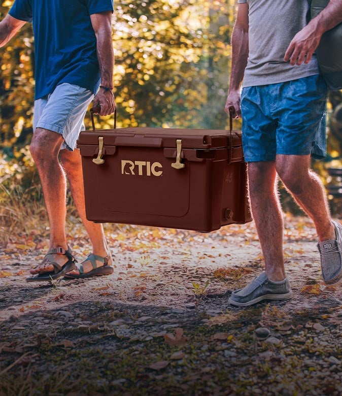 Two men carrying a brown RTIC hard-sided cooler by its side handles while walking outdoors on a wooded trail.