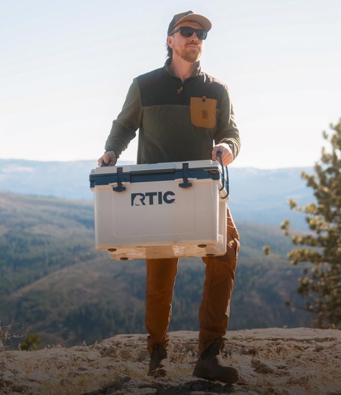 Hiker carrying a hardsided RTIC cooler on a rocky overlook with mountains in the background