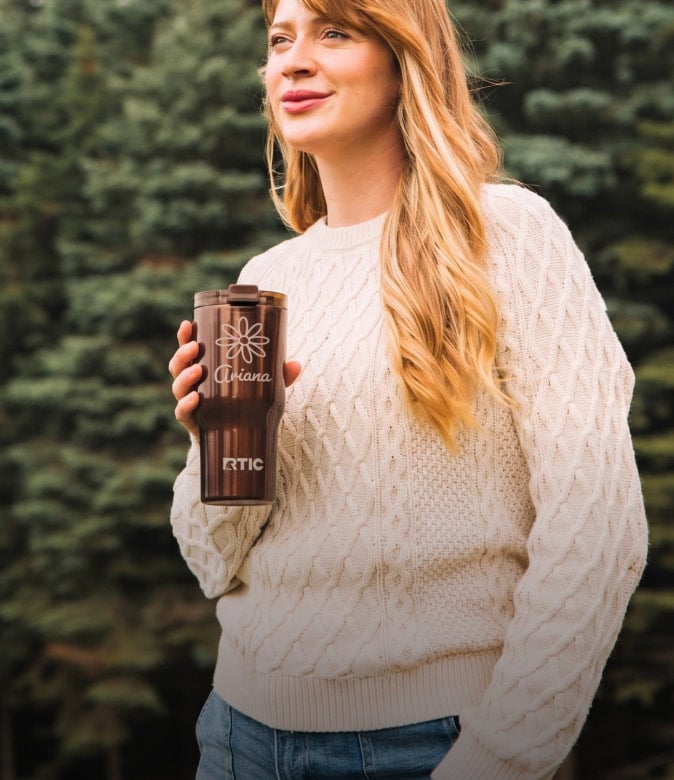 Woman in a cream sweater holding a personalized brown RTIC tumbler outdoors.