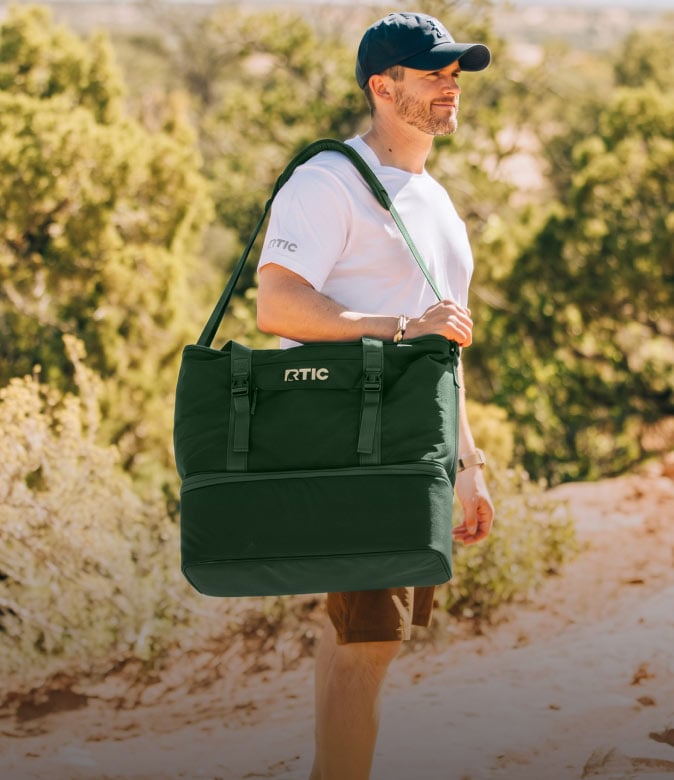 Man carrying a large green RTIC insulated cooler tote along a sunlit outdoor trail.