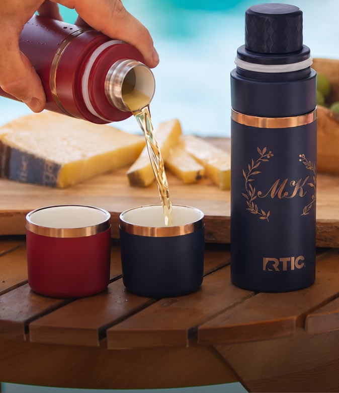 RTIC insulated flask pouring a cold drink into two shot glasses beside food on a wooden table.
