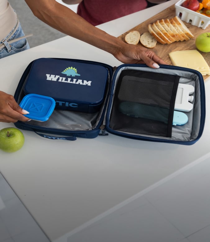 Open lunch bag with a food container labeled “William,” alongside bread, cheese, and apples on a kitchen table.