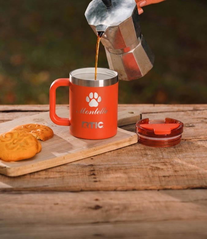 Coffee pouring from a coffee pot into an orange RTIC travel mug customized with a paw print logo, next to two cookies.