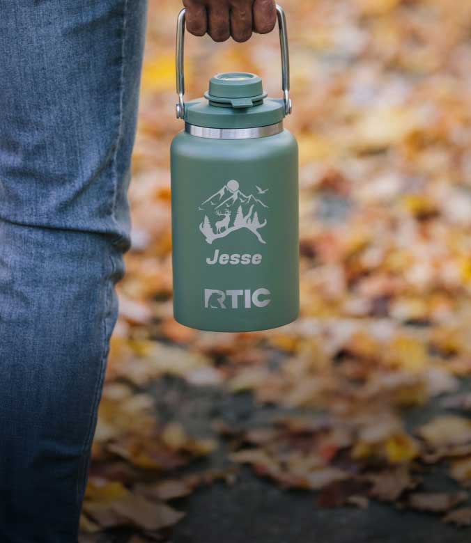 A close-up of a person holding a sage green RTIC jug engraved with mountains, wildlife, and the name Jesse.