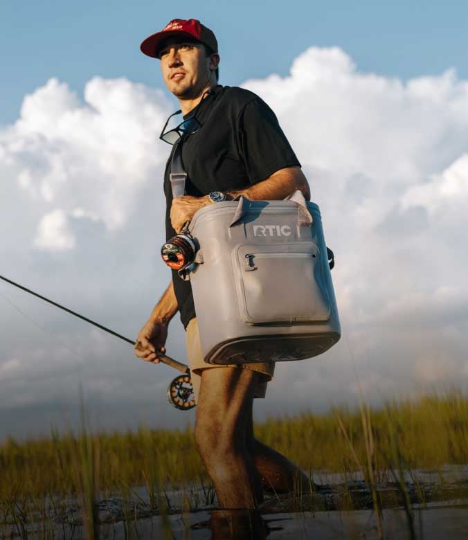 A man walks through a grassy marsh carrying a grey RTIC soft cooler over his shoulder and holding a fishing rod.
