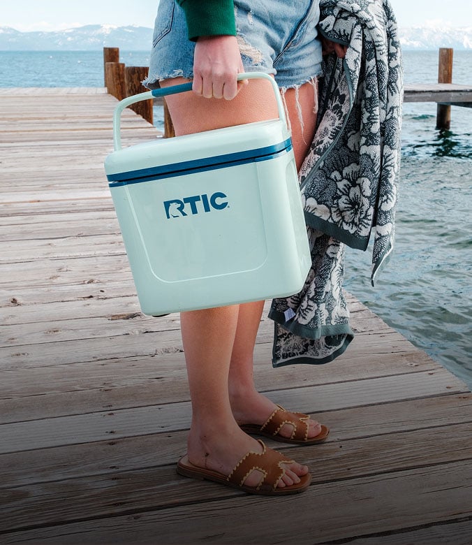 A close-up of a person standing on a dock, holding a small teal RTIC hard cooler with a handle.