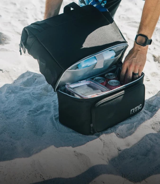 Open black RTIC cooler backpack filled with food and drinks on a sandy beach.