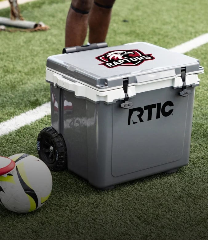 Gray RTIC wheeled cooler with a Raptors logo beside soccer balls on a turf field.