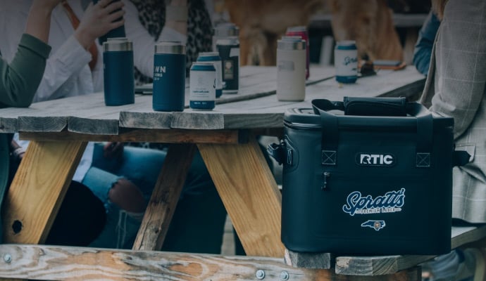 A black RTIC soft cooler customized with a customer logo on the front pocket; sitting on a bench.