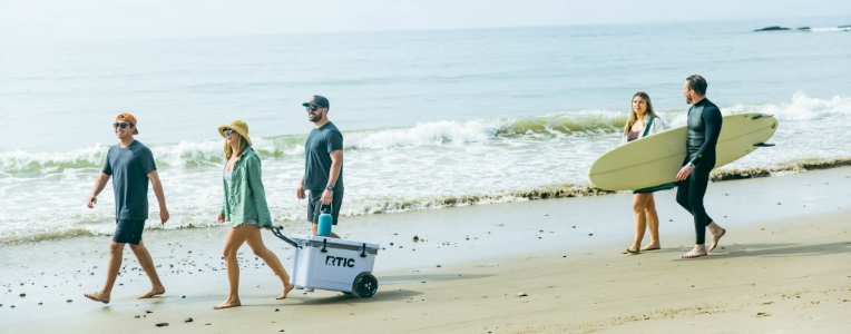 A group of friends walking with an RTIC wheeled cooler and a surfboard at the beach.