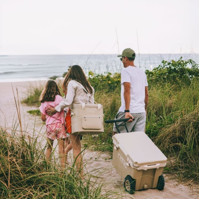 A family walking to the beach with a tan RTIC wheeled hard cooler and tan RTIC soft cooler.