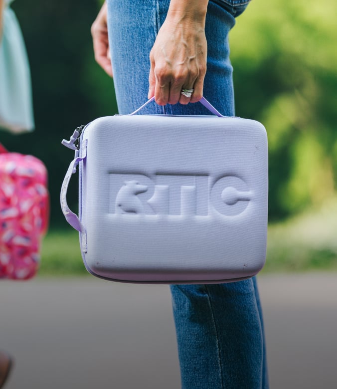 Person holding a lavender RTIC cooler lunch box outdoors, wearing jeans with a blurred green background.