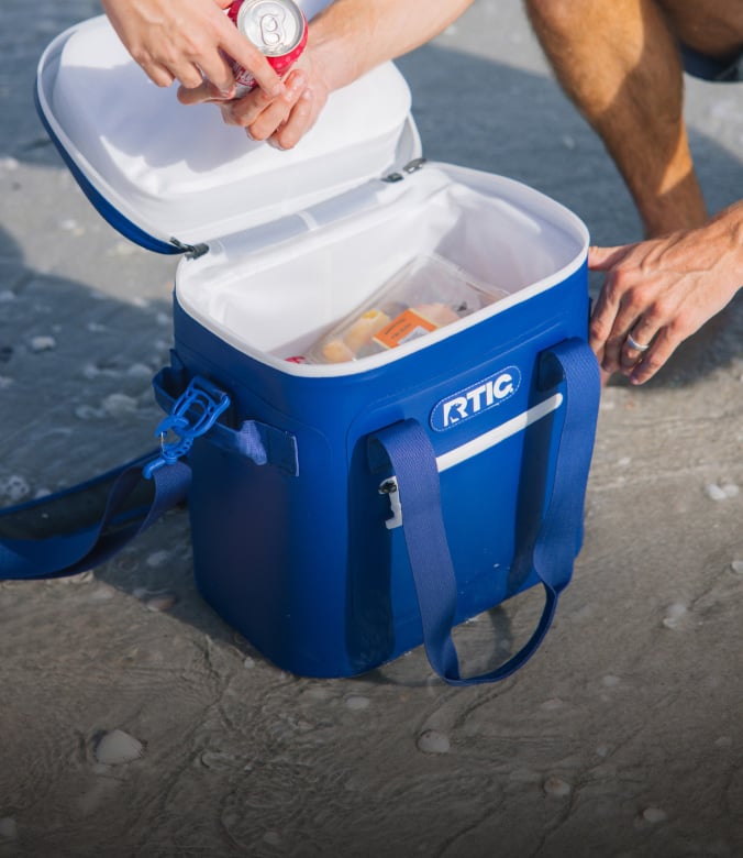 Blue RTIC soft cooler open on a beach shoreline as people place canned drinks and snacks inside.