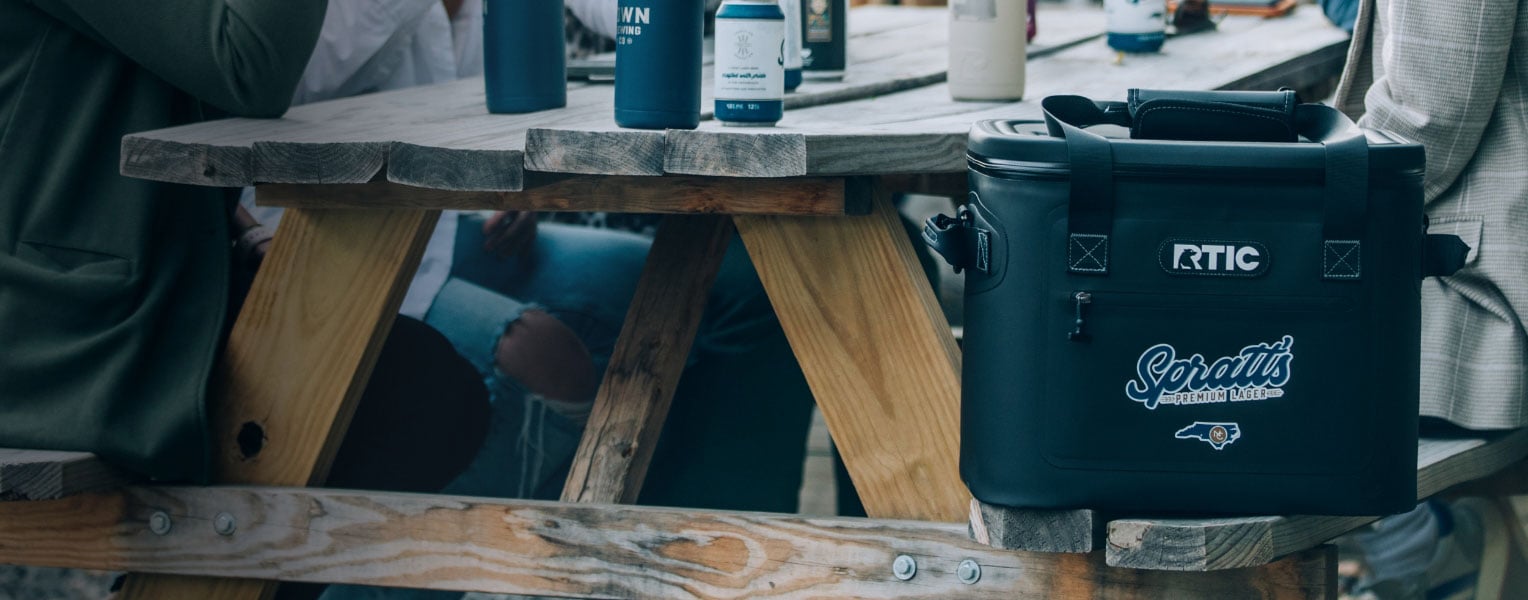 A black RTIC soft cooler featuring a customer logo on the front pocket sitting on a table.