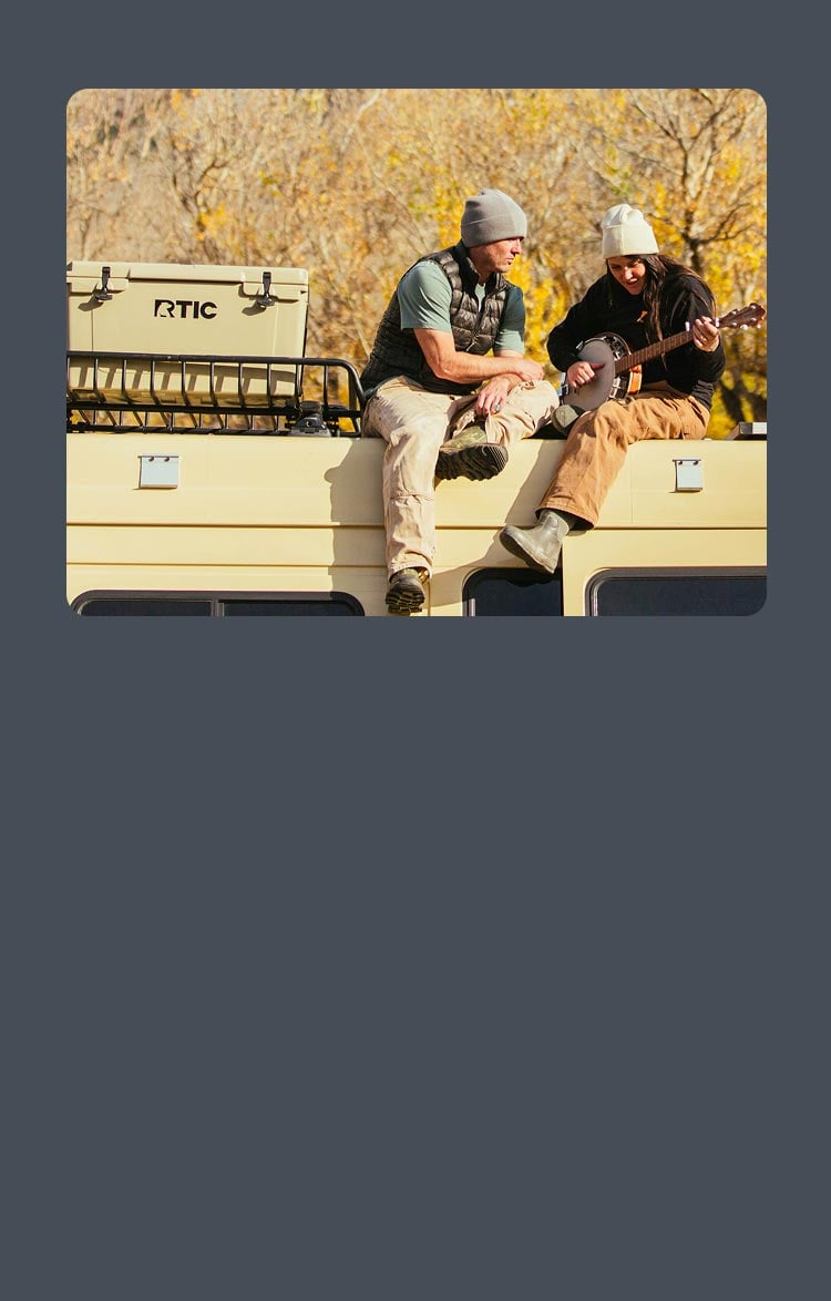 Two people sitting on the roof of a van, next to a RTIC Cooler; one is playing a banjo.