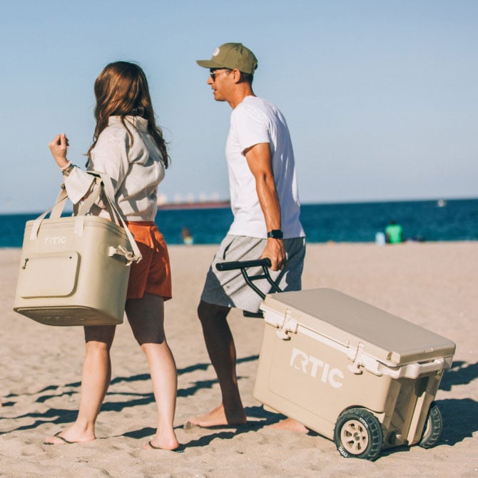 Two people walk on a sandy beach carrying a cooler bag and a wheeled cooler, with the ocean visible in the background.
