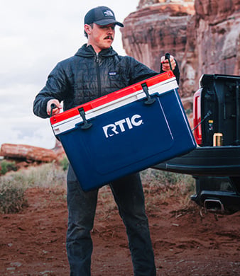 Man carrying a large red, white and blue RTIC hard cooler beside a pickup truck in a rocky desert landscape.