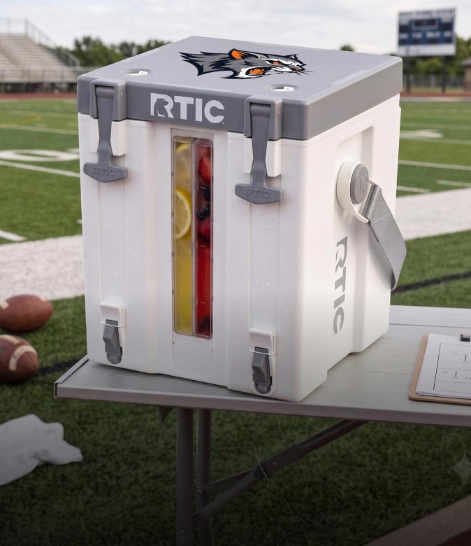 Customized RTIC water cooler with team logo, gray lid, and clear front panel displayed on a sports field for outdoor hydration.