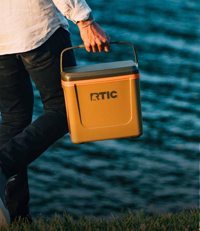 Person carrying a tan RTIC hard cooler by the water at sunset, showcasing a portable insulated cooler for outdoor adventures.