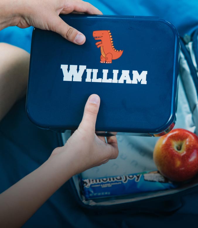 Personalized navy lunch box with the name William and a dinosaur graphic placed inside a backpack with an apple and snacks.